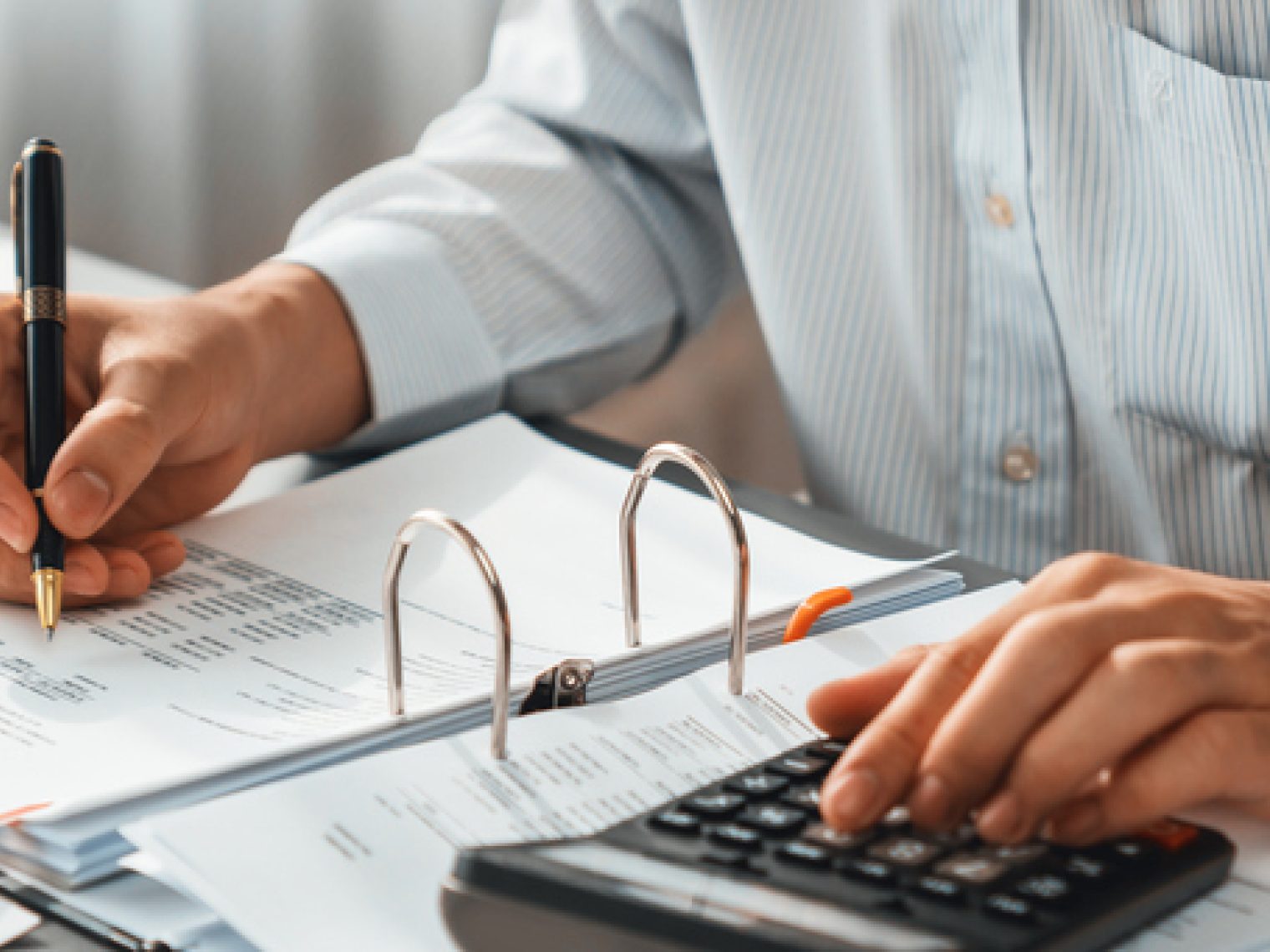 A person is working at a desk with financial documents, using a calculator with one hand and holding a pen in the other.