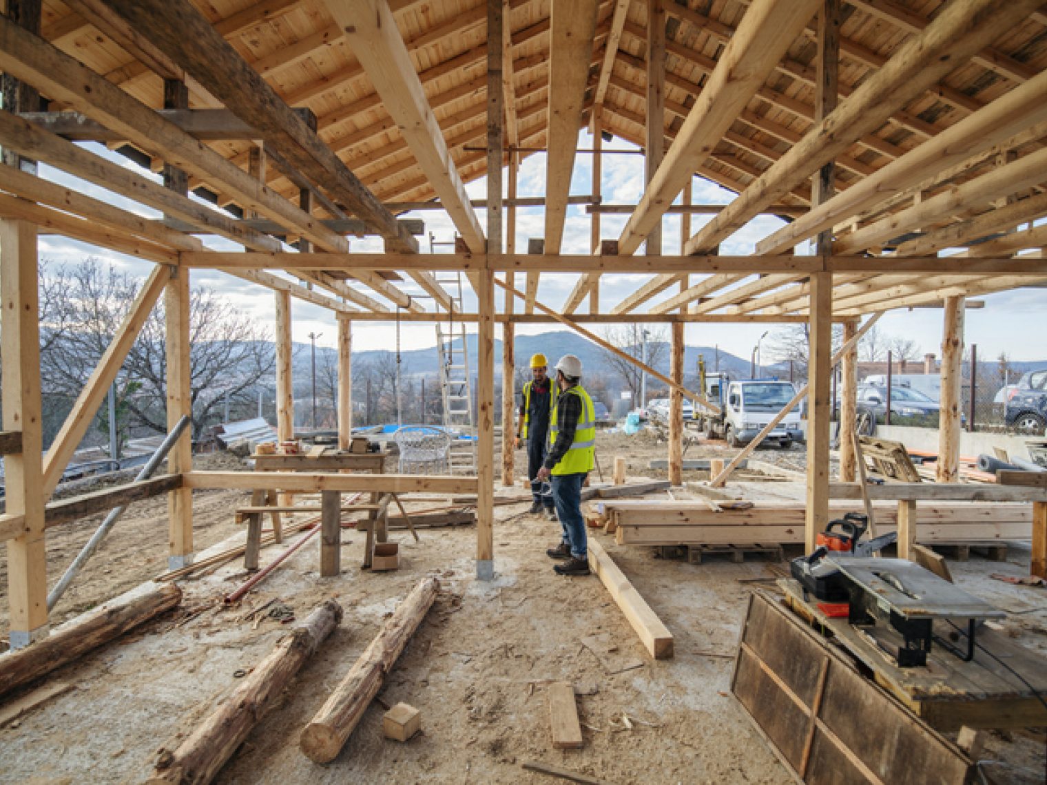 Two construction workers in high visibility jackets are discussing plans inside the wooden frame of a house under construction, surrounded by tools and building materials.