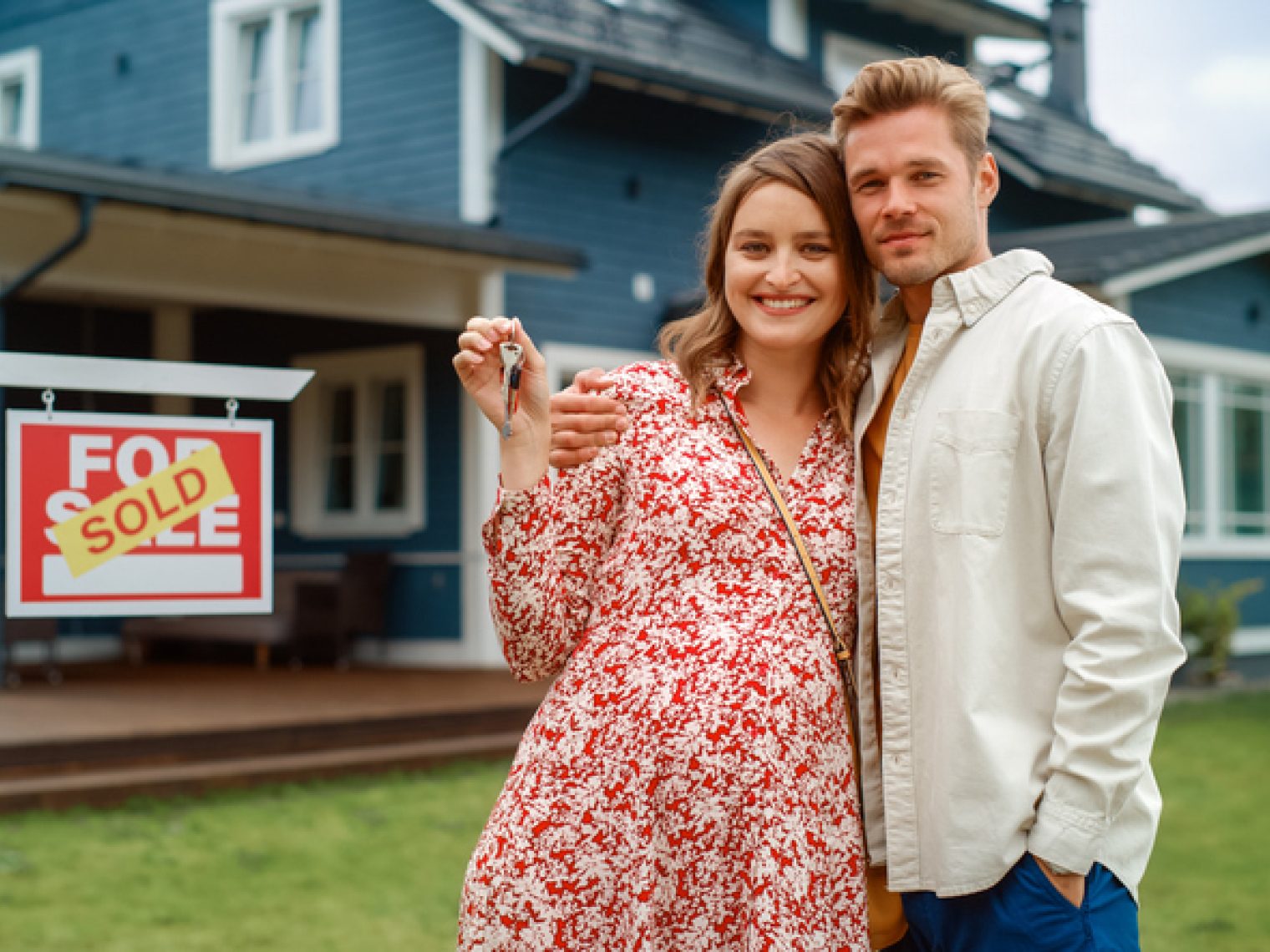 A happy couple embraces and smiles, holding a set of keys in front of a blue house with a "Sold" sign, celebrating their new home purchase.