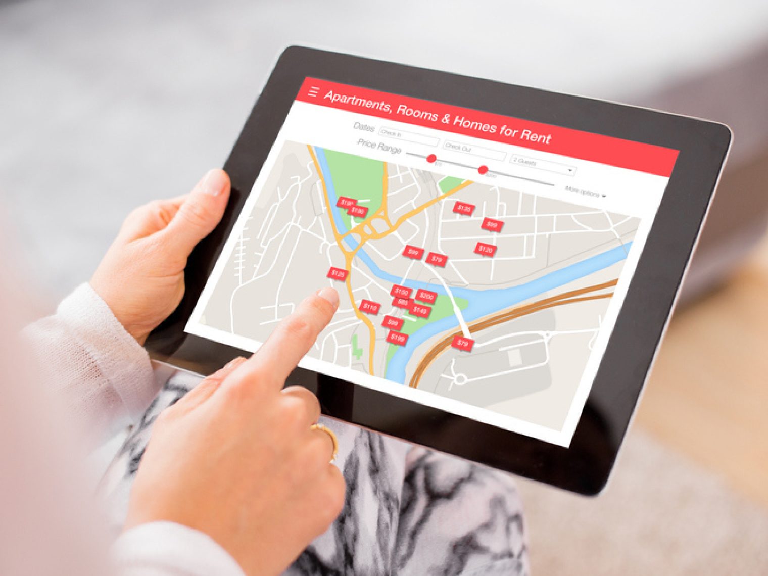 Close-up of a person browsing a real estate rental app on a tablet, with a map displaying various listings indicated by price tags in a specific area.