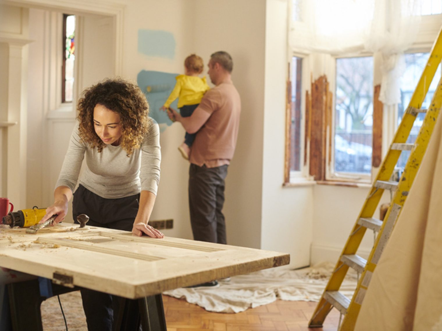 A woman is focused on a DIY woodworking project on a sturdy table in a room that appears to be undergoing renovations. In the background, a man is holding a baby, standing next to a yellow ladder and painting the wall with a roller.