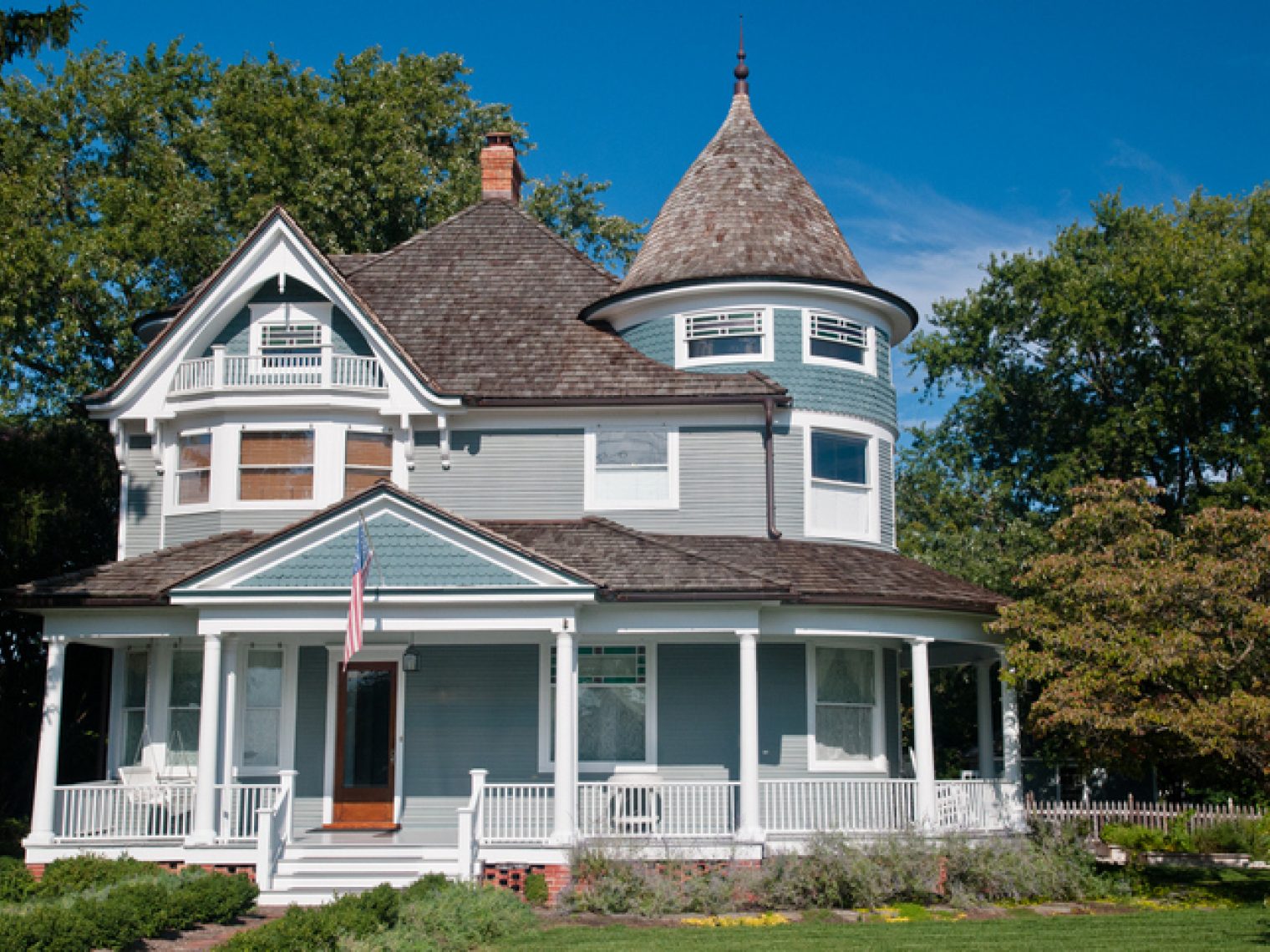 A Victorian-style house with a distinctive turret and wrap-around porch, set against a clear blue sky with surrounding greenery, exhibiting classic American architectural elements.