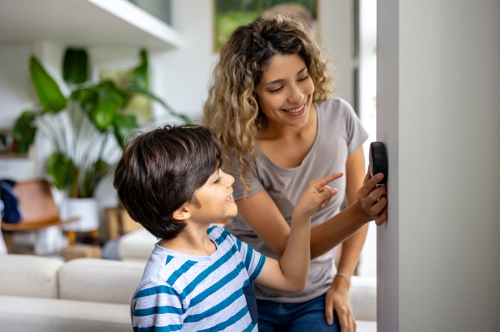 A smiling young boy in a striped blue and white T-shirt looks up at a woman who is pointing to a digital thermostat on the wall.