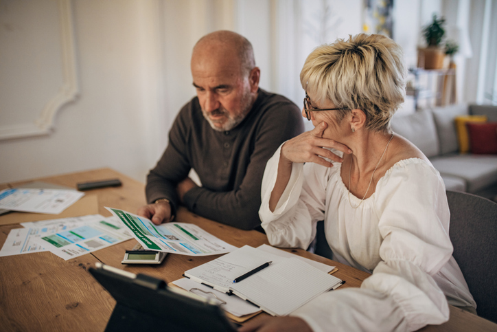 An older couple appears concerned while reviewing financial documents spread out on a table.