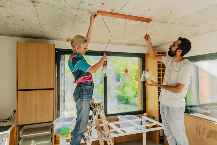 A woman and a man are working together to install a wooden light fixture with three hanging bulbs in a room with a modern aesthetic. The woman is standing on a stepladder, securing the fixture to the ceiling, while the man is holding it in place, providing support and guidance.